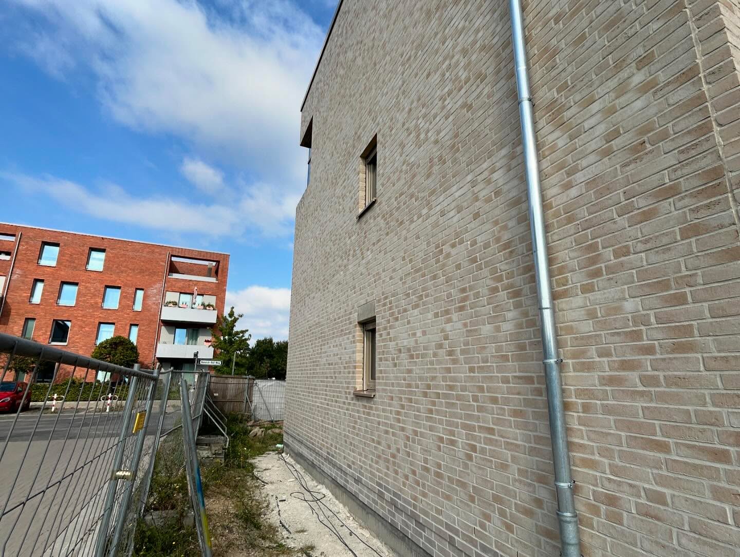 Gebäudeecke in hellem Klinker mit Fallrohr und rotem Nachbargebäude